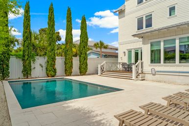 Inviting turquoise backyard pool beside a white coastal-style house, sunlit stone patio with lounge chairs and tall cypress trees.