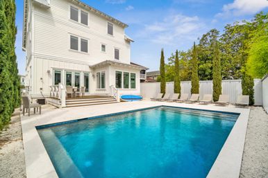Luxury white three-story house with a rectangular blue backyard swimming pool, poolside loungers, tall cypress trees and a small deck dining area