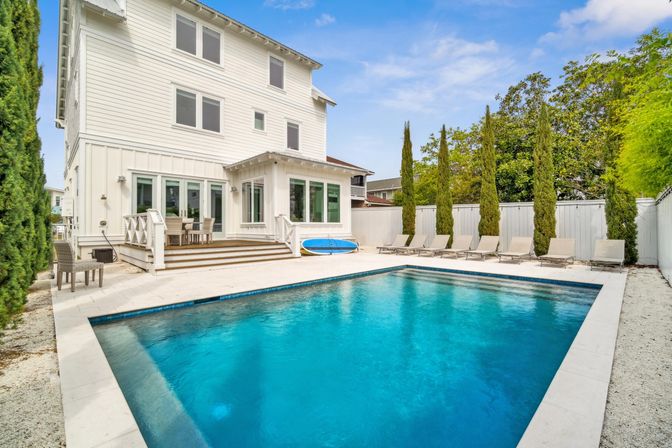 Luxury white three-story house with a rectangular blue backyard swimming pool, poolside loungers, tall cypress trees and a small deck dining area