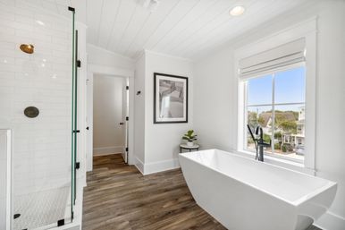 Bright modern bathroom with freestanding white tub and black floor-mounted faucet, glass-enclosed shower with white subway tile, wood-look flooring and a large window overlooking neighboring homes.