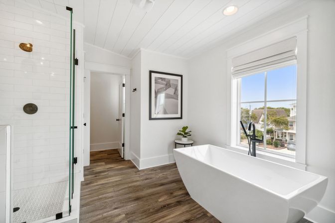 Bright modern bathroom with freestanding white tub and black floor-mounted faucet, glass-enclosed shower with white subway tile, wood-look flooring and a large window overlooking neighboring homes.