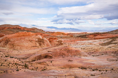 Expansive red‑rock desert with layered sandstone formations, sparse desert scrub and rolling hills stretching to a distant mountain range under a dramatic cloudy sky — Southwestern US arid landscape.