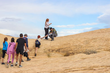 Group of hikers climbing a sloped sandstone rock formation under a bright blue sky; one hiker reaches down to help another up while others wait below, backpacks and casual outdoor gear visible.