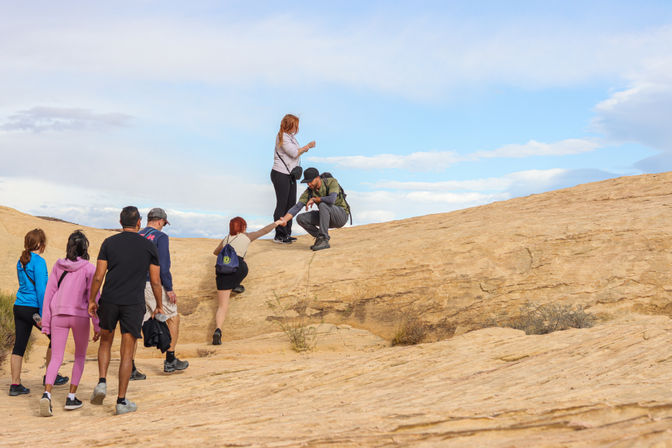 Group of hikers climbing a sloped sandstone rock formation under a bright blue sky; one hiker reaches down to help another up while others wait below, backpacks and casual outdoor gear visible.
