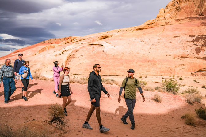 Group of hikers on a red‑rock desert day hike, walking past smooth sandstone cliffs under a dramatic cloudy sky