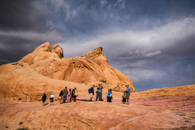Group of hikers traversing pink-orange sandstone slopes beneath rounded red rock formations and a dramatic cloudy sky in a Southwestern desert