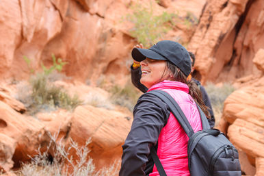 Smiling hiker in a black cap and bright pink puffer vest with a backpack exploring a red sandstone canyon with desert shrubs — red rock canyon hiking scene