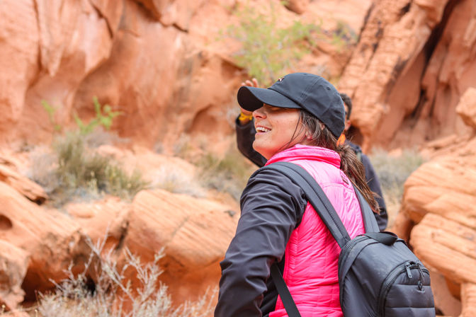Smiling hiker in a black cap and bright pink puffer vest with a backpack exploring a red sandstone canyon with desert shrubs — red rock canyon hiking scene