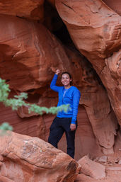 Smiling hiker in a bright blue jacket striking a playful pose inside a red sandstone canyon alcove, surrounded by layered desert red rocks with a blurred green shrub in the foreground.