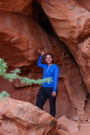 Smiling hiker in a bright blue jacket striking a playful pose inside a red sandstone canyon alcove, surrounded by layered desert red rocks with a blurred green shrub in the foreground.