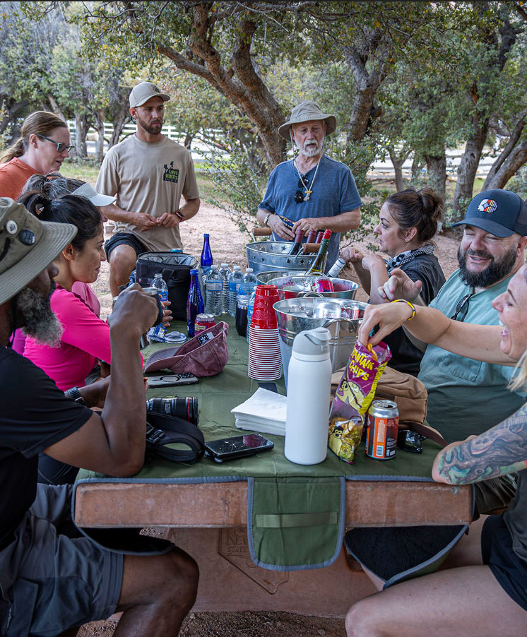 Group enjoying an outdoor picnic at a shaded campground table — friends chatting around snacks, bottled water, red cups and beverage buckets under trees