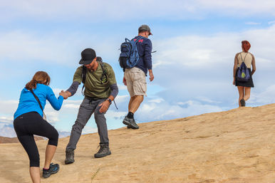 Four hikers with backpacks ascending a smooth sandstone slope — one offers a helping hand to another — under a bright blue sky in a desert hiking scene.