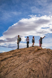 Five hikers standing on a rocky sandstone outcrop at a scenic overlook, one taking a selfie against a dramatic blue sky with layered clouds — outdoor adventure in a desert rock landscape.