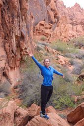 Smiling hiker in a blue jacket and hiking shoes standing on a red sandstone boulder with arms outstretched amid red rock canyon and desert shrubs, American Southwest hiking scene.