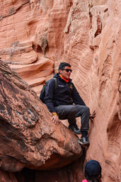 Hiker in sunglasses and backpack squeezes into a narrow red sandstone slot canyon, sitting on a ledge while rock-scrambling in outdoor gear — red rock adventure scene.