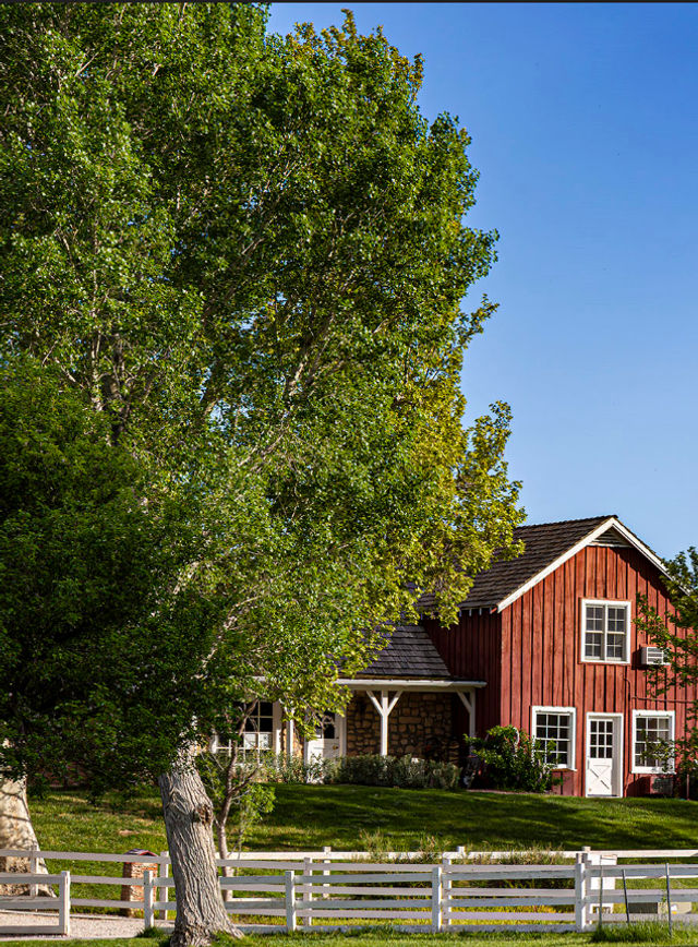 Red wooden barn-style house with white-trim windows and door, partially hidden by tall leafy trees, white fence and green lawn under a clear blue sky in a rural farmhouse setting