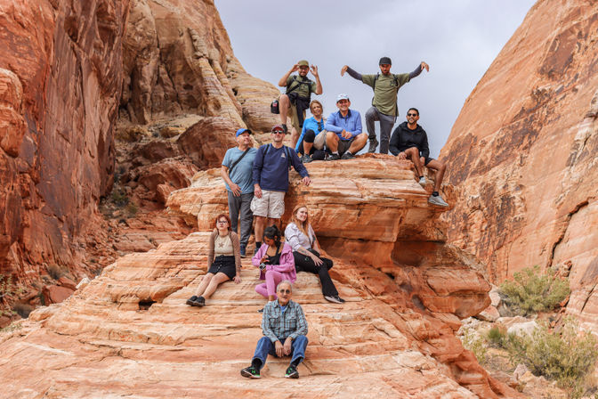 Group of hikers sitting and standing on layered red sandstone outcrop in a southwestern desert canyon under a cloudy sky