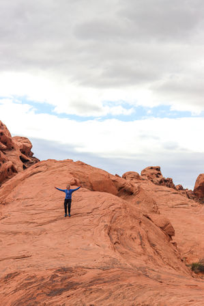 Hiker in a blue jacket standing arms outstretched on red sandstone rock formations under a cloudy sky in a southwestern desert landscape.