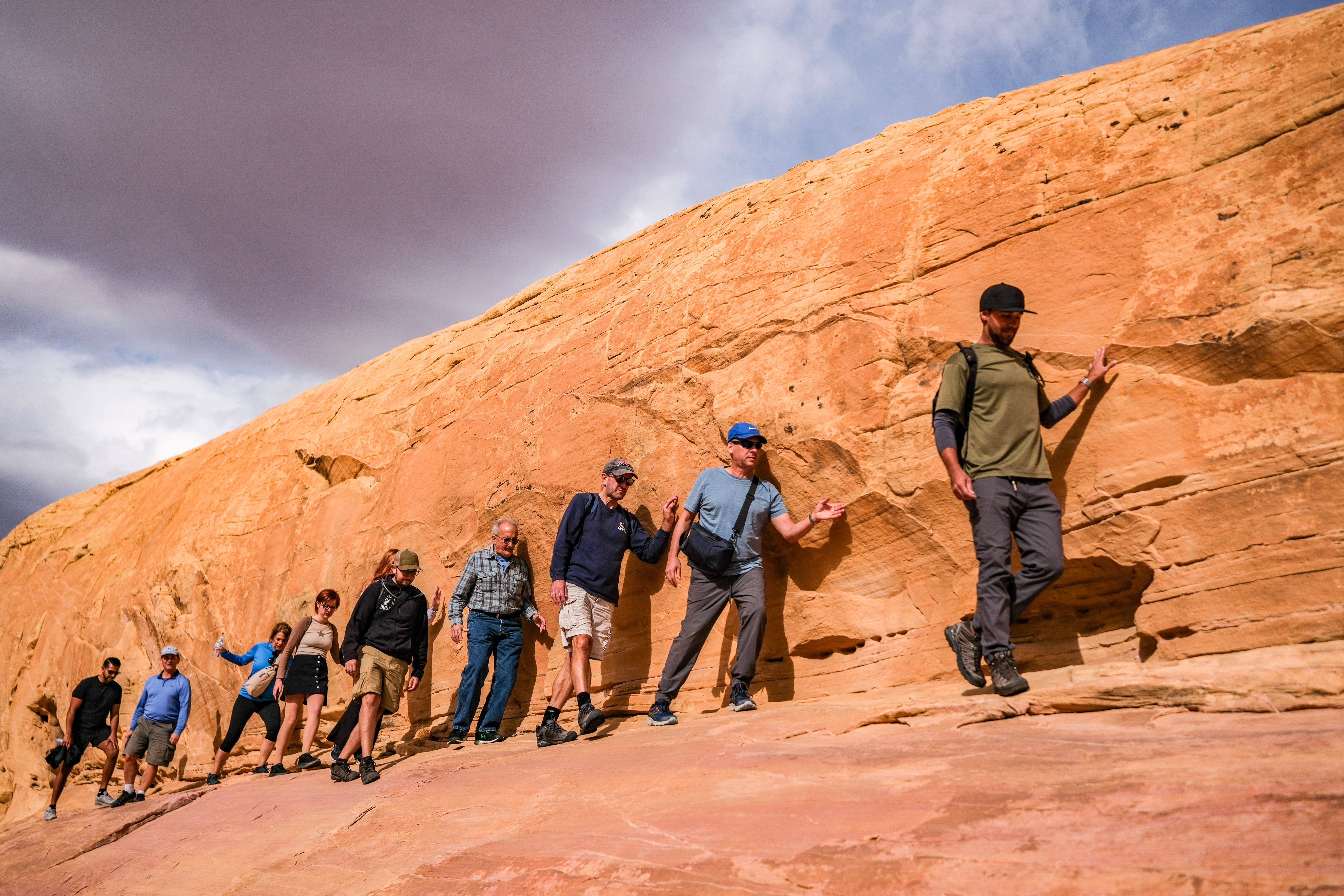 Group of hikers walking single-file along a sloping red sandstone cliff in the American Southwest, using the rock for balance beneath a dramatic cloudy sky