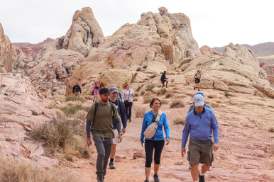 Group of hikers on a scenic red sandstone desert trail winding through layered pink-and-cream rock formations and sparse desert shrubs.
