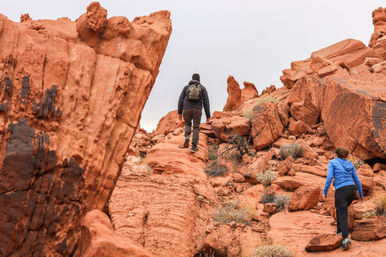 Two hikers climbing a red sandstone slope among towering orange rock formations and sparse desert scrub under an overcast sky — red rock hiking adventure.