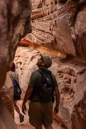 Hikers exploring a narrow red sandstone slot canyon, man with backpack and cap looking up at layered red rock walls