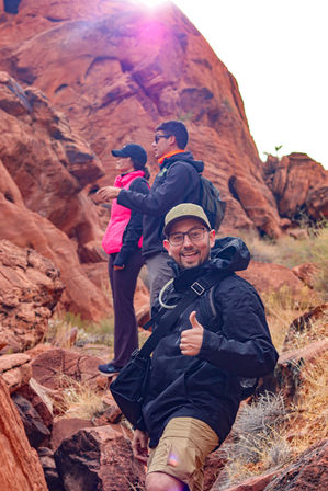 Smiling hiker giving a thumbs-up in the foreground with two companions hiking a red sandstone canyon trail under a sun flare