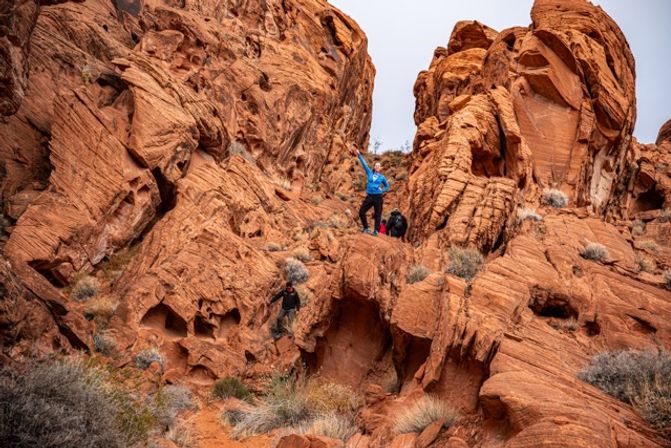 Two hikers exploring towering red sandstone canyon walls — one in a blue jacket waving from a ledge while the other climbs among sculpted rocks and desert scrub.