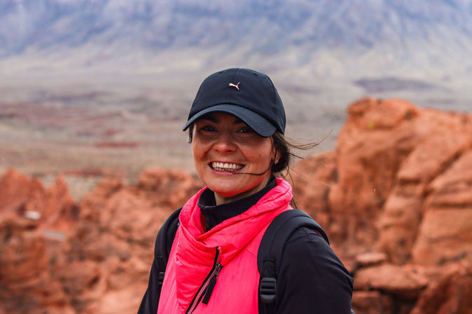 Smiling hiker wearing a black cap and bright pink vest with backpack on red-rock desert cliffs, scenic red-rock canyon landscape and distant mountains