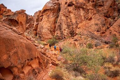 Small group of hikers trekking a winding red sandstone canyon trail amid dramatic rock formations, sparse desert shrubs and rugged cliffs under a cloudy sky