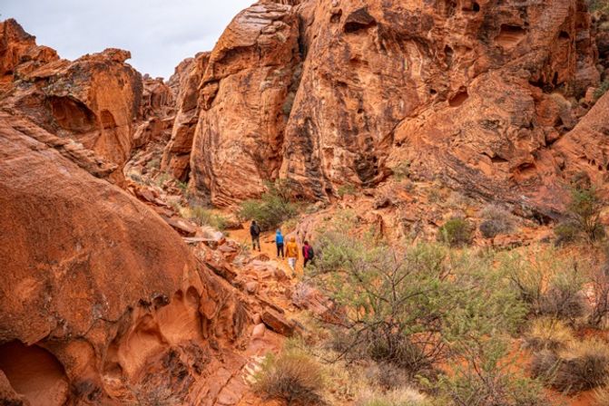 Small group of hikers trekking a winding red sandstone canyon trail amid dramatic rock formations, sparse desert shrubs and rugged cliffs under a cloudy sky