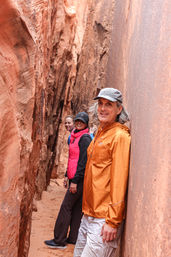 Three hikers squeeze through a narrow red sandstone slot canyon with a sandy floor — man in an orange jacket leads, woman in a pink vest and black cap follows — scenic American Southwest slot-canyon hike