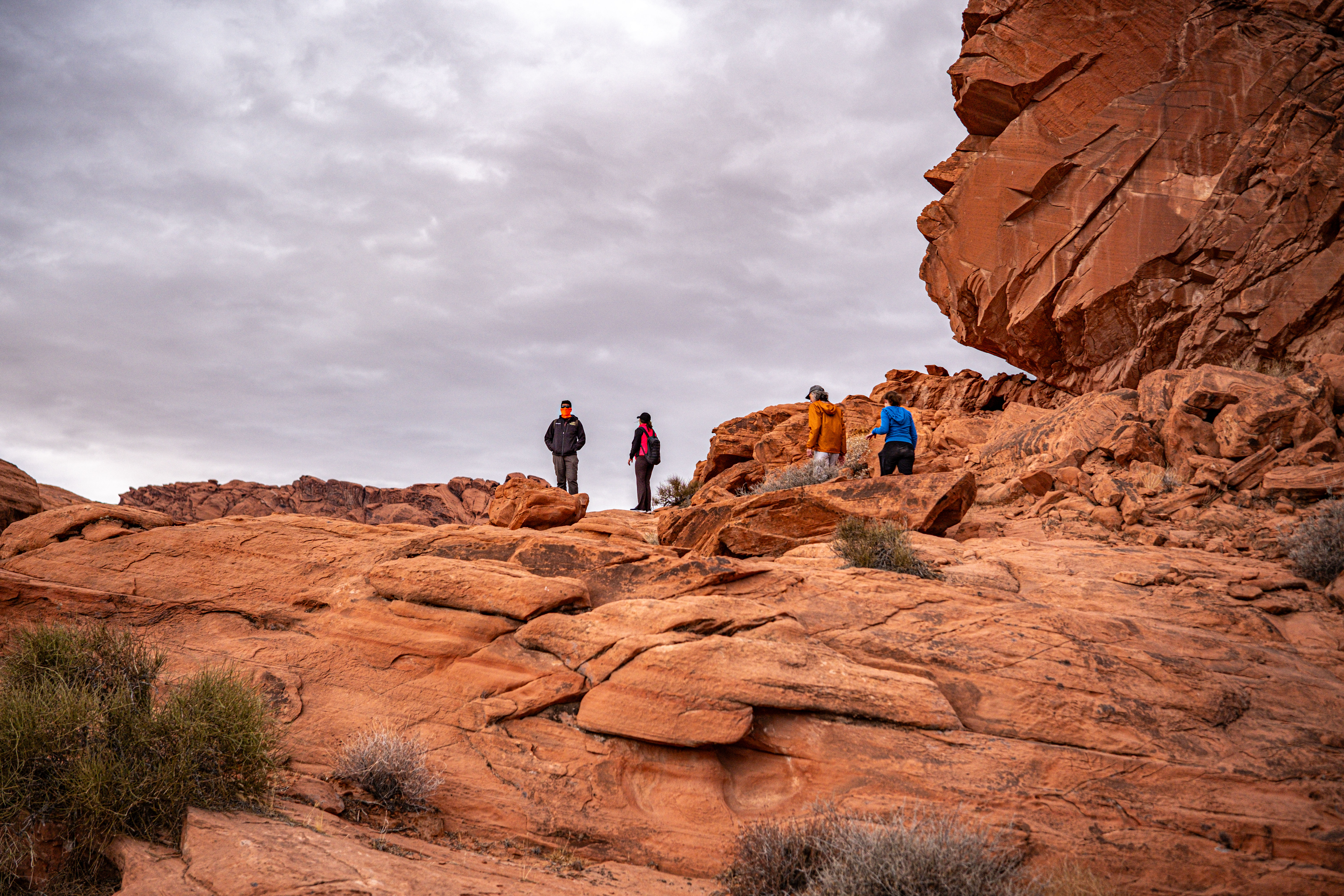 Four hikers exploring orange-red sandstone cliffs under an overcast sky in a scenic Southwestern desert rock formation
