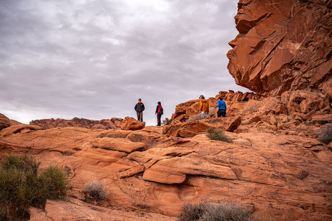 Four hikers exploring orange-red sandstone cliffs under an overcast sky in a scenic Southwestern desert rock formation