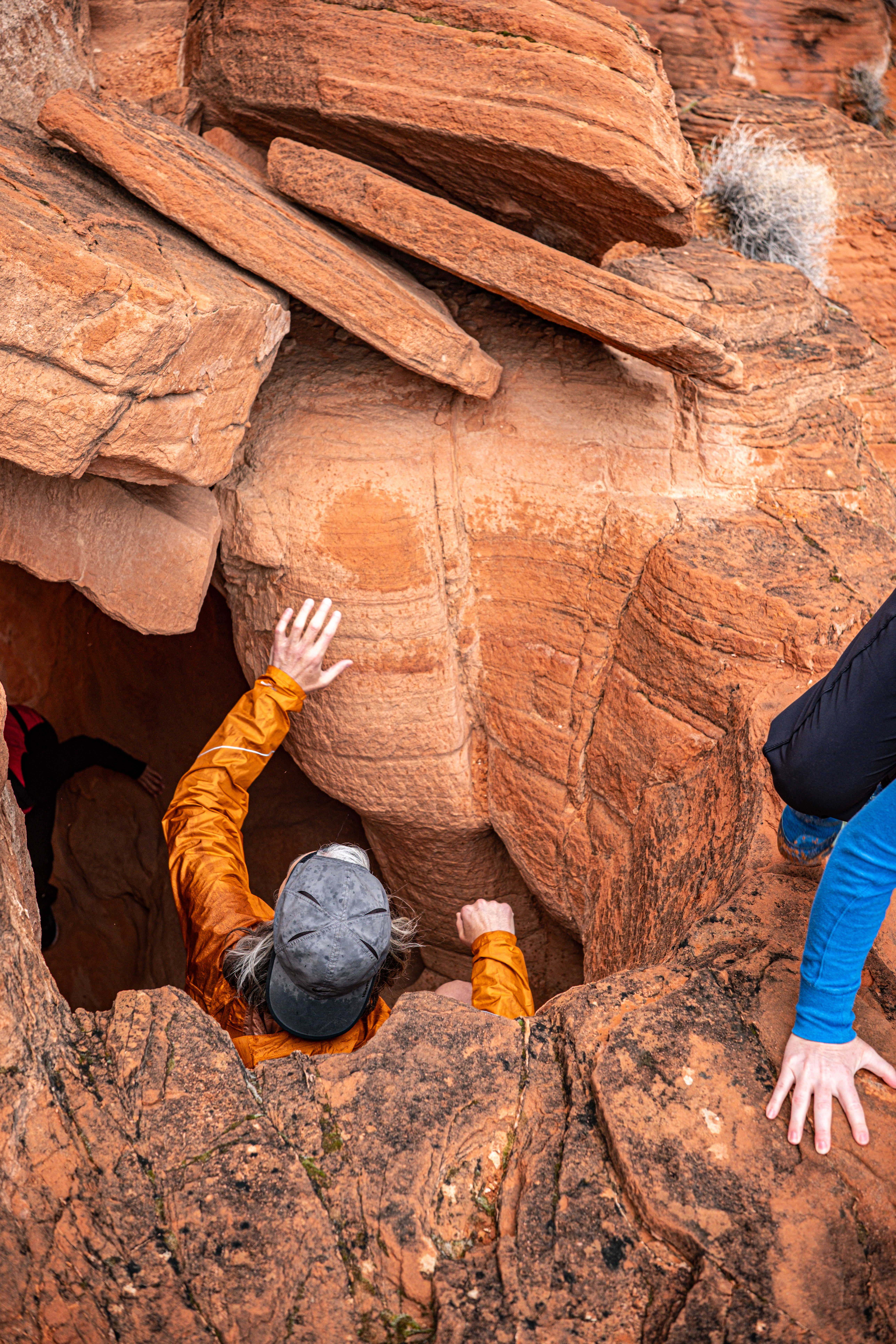 Hiker in an orange jacket and gray cap climbing down a narrow red sandstone slot canyon, hands gripping layered rock while a companion's hand in a blue sleeve reaches at the rim — desert red-rock adventure.