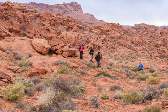 Small group of hikers traversing red sandstone desert with rugged rock formations and sparse scrub under an overcast sky