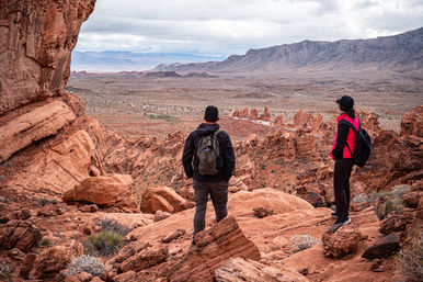 Two hikers with backpacks on red sandstone cliffs overlooking a sweeping red‑rock desert valley, jagged rock formations and distant mountains beneath a moody cloudy sky