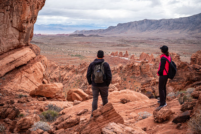 Two hikers with backpacks on red sandstone cliffs overlooking a sweeping red‑rock desert valley, jagged rock formations and distant mountains beneath a moody cloudy sky