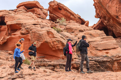 Five hikers traversing layered red sandstone formations in a desert canyon, wearing bright outdoor gear and backpacks under a cloudy sky.