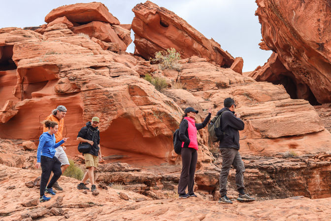 Five hikers traversing layered red sandstone formations in a desert canyon, wearing bright outdoor gear and backpacks under a cloudy sky.