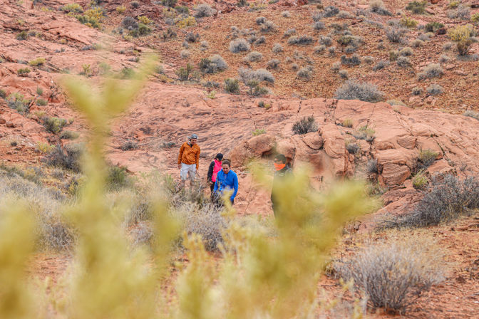 Group of hikers in colorful jackets walking through a red sandstone canyon with sparse desert scrub and rocky trails