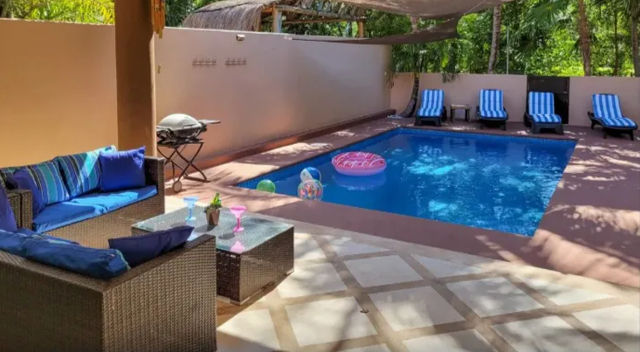 Tropical backyard patio with a rectangular blue swimming pool, striped lounge chairs, wicker outdoor sofa with blue cushions, inflatable float and beach balls in the water, shaded canopy and small barbecue grill.
