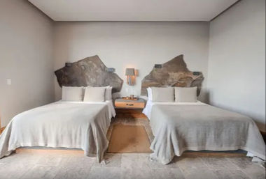 Contemporary hotel room with two beds in light gray linens, sculpted natural stone headboards, wooden nightstand with lamp between them, neutral walls and tiled floor — minimalist boutique style.