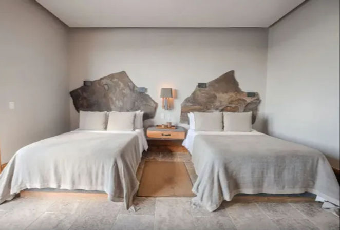 Contemporary hotel room with two beds in light gray linens, sculpted natural stone headboards, wooden nightstand with lamp between them, neutral walls and tiled floor — minimalist boutique style.