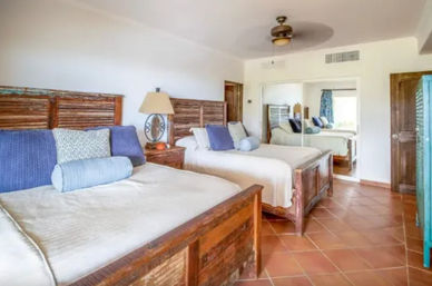 Sunny coastal-style twin bedroom with rustic reclaimed-wood beds, blue accent pillows and bolsters, terracotta tile floor, ceiling fan, mirrored closet and bedside lamp