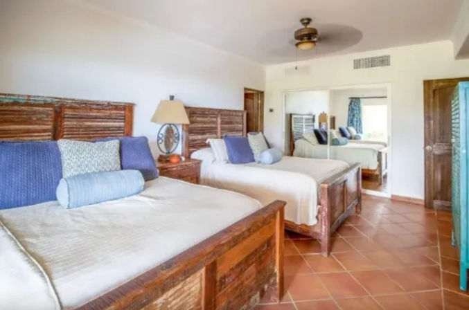 Sunny coastal-style twin bedroom with rustic reclaimed-wood beds, blue accent pillows and bolsters, terracotta tile floor, ceiling fan, mirrored closet and bedside lamp