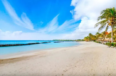 Sunny tropical white-sand beach with turquoise water, palm-lined shoreline, small resort huts and wispy clouds in a bright blue sky.