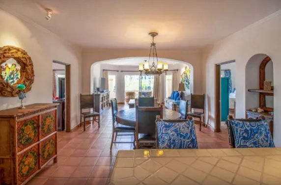 Mediterranean-style open-plan dining and living room with terracotta tile floor, arched doorways, round wooden dining table, blue paisley upholstered chairs, decorative painted chest, chandelier and sunlit seating area by sliding glass doors.