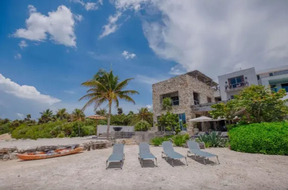 Four sun loungers on a sandy tropical beach in front of a stone beachfront villa with a palm tree and kayak under a bright blue sky