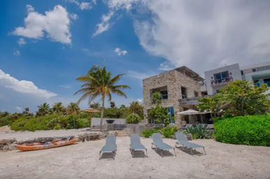Four sun loungers on a sandy tropical beach in front of a stone beachfront villa with a palm tree and kayak under a bright blue sky